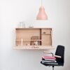 A minimalist, wall-mounted wooden desk is open, revealing internal compartments and various desk items, with a black chair and pink pendant light