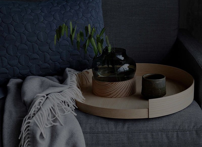 gray sofa featuring a blue cushion, a light-colored throw blanket, a round wooden tray holding a glass Ikebana vase with green sprigs, and a small dark cup.